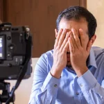 a man sitting on a sofa with his head in his hands while a camera on a tripod films him