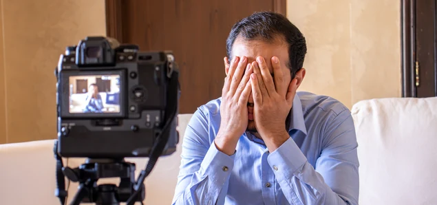 a man sitting on a sofa with his head in his hands while a camera on a tripod films him