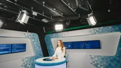 a news presenter at the desk of a small TV studio under studio lights