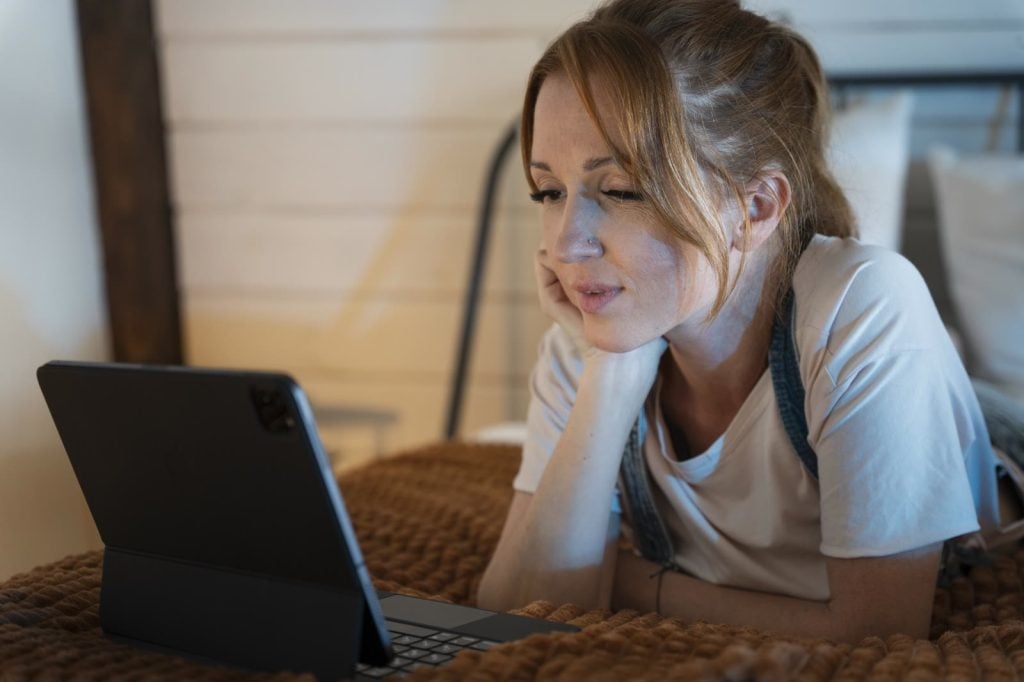 A woman lies on a bed and watches a stream on her tablet, chin on her hand