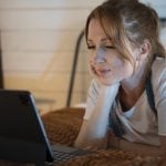 A woman lies on a bed and watches a stream on her tablet, chin on her hand