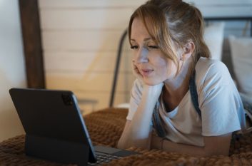 A woman lies on a bed and watches a stream on her tablet, chin on her hand