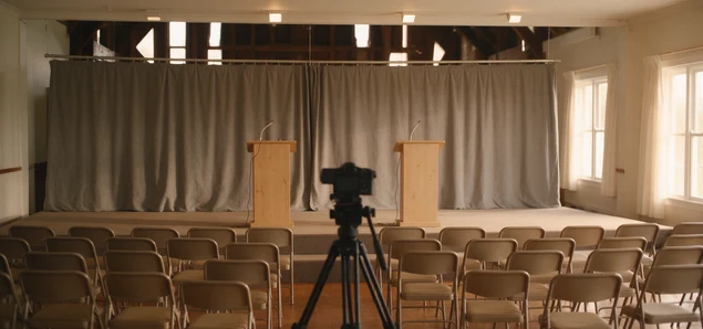 two empty wooden lecterns on a small community hall stage, a video camera on a tripod facing them over rows of folding chairs